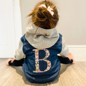 May include: A denim jacket with a gray hood and sleeves. The back of the jacket features a floral embroidered letter "B" in pink, yellow, and purple. The jacket is worn by a child.
