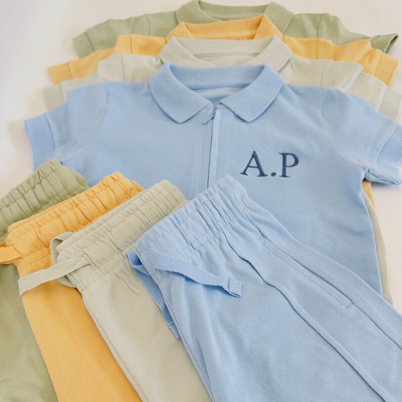 May include: A stack of light blue, yellow, and green clothing items. The top item is a light blue polo shirt with a zipper and the embroidered initials "A.P.". The bottom items are a stack of light blue, yellow, and green pants.
