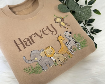 May include: A tan sweatshirt with an embroidered design of a lion, elephant, giraffe, zebra, and monkey. The name "Harvey" is embroidered above the animals.