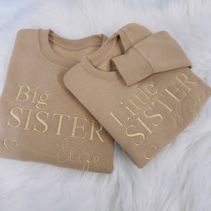 May include: Two tan sweatshirts with gold embroidery. The larger sweatshirt says "Big Sister" with the name "Eliza" underneath. The smaller sweatshirt says "Little Sister" with the name "Elyana" underneath.