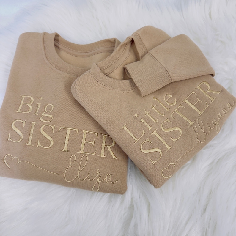 May include: Two tan sweatshirts with gold embroidery. The larger sweatshirt says "Big Sister" with the name "Eliza" underneath. The smaller sweatshirt says "Little Sister" with the name "Elyana" underneath.