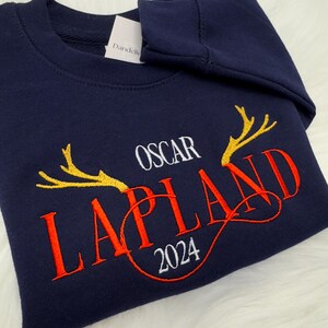 May include: A navy blue sweatshirt featuring embroidered text and reindeer antlers. The word "OSCAR" is above "LAPLAND" in red, with "2024" below. Gold antlers flank "LAPLAND". A white tag is visible.