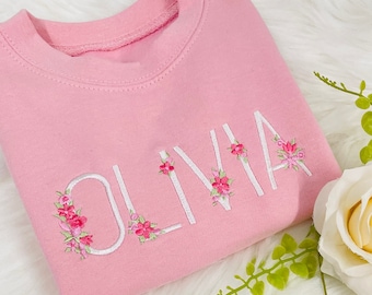 May include: Pink sweatshirt with the name "Olivia" embroidered in white with pink floral accents.