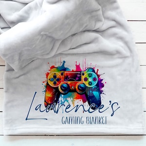 May include: A pale grey blanket featuring a vibrant, watercolour-style video game controller design. The controller is in rainbow hues. Below, the text reads "Lawrence's GAMING BLANKET" in blue script.