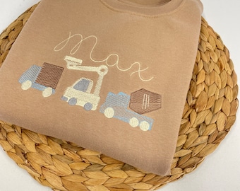 Puede incluir: Una sudadera marrón con el nombre "Max" bordado en la parte delantera. La sudadera presenta un diseño de un camión de construcción, un camión volquete y una caja.