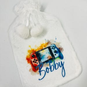 May include: White fuzzy hot water bottle with a watercolor design of a Nintendo Switch controller and the name "Bobby" printed on it.
