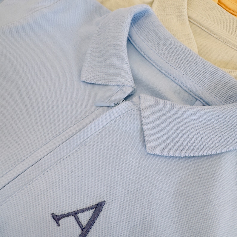 May include: A light blue polo shirt with a zipper closure and a navy blue embroidered letter 'A' on the front.