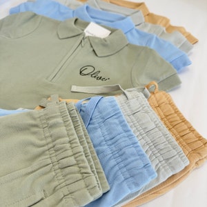 May include: A stack of five different colored pants with a green polo shirt on top. The polo shirt has the word "Olive" embroidered on the left side of the chest.