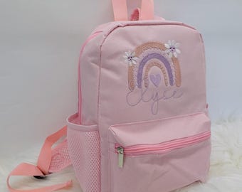 May include: Pink canvas backpack with a rainbow and flower embroidery. The name "Lyse" is embroidered below the rainbow.