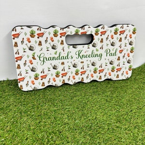 May include: A rectangular kneeling pad featuring a white background with a pattern of gardening tools and plants. It has a black border and a handle. The text "Grandad's Kneeling Pad" is written in green script.