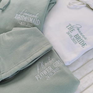 May include: Three hooded sweatshirts with embroidered text. The first sweatshirt is sage green with the text "Hannah Bridesmaid 24.05.2025". The second sweatshirt is white with the text "Joanna The Bride 24.05.2025". The third sweatshirt is sage green with the text "Phoebe Flower Girl 24.05.2025".