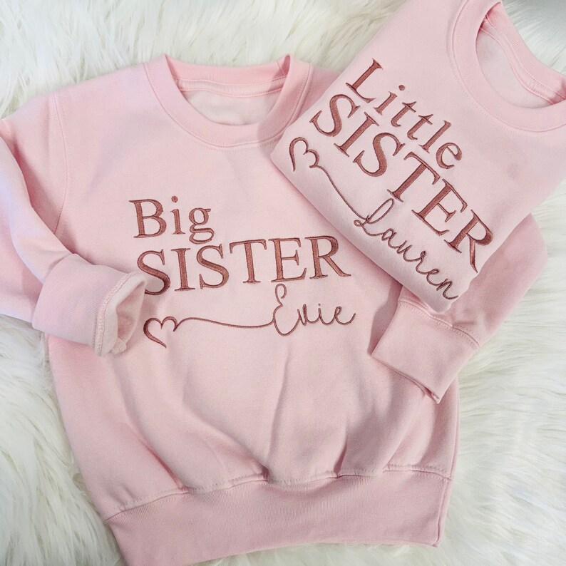 May include: Two pink sweatshirts with the text "Big Sister & Evie" and "Little Sister Lauren" in rose gold lettering.
