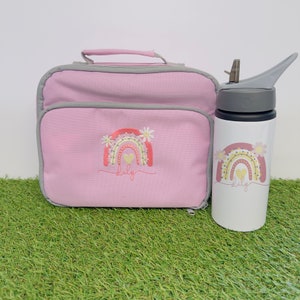 May include: A pink lunch bag and water bottle set. The lunch bag has a grey trim and a rainbow design with the name "Lily" embroidered on the front. The white water bottle features a matching rainbow design and the name "Lily".