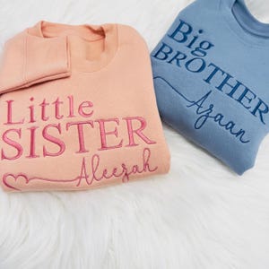 May include: Two sweatshirts, one peach and one blue, with embroidered text. The peach sweatshirt says "Little Sister Aleezah" with a pink heart. The blue sweatshirt says "Big Brother Ayaan".