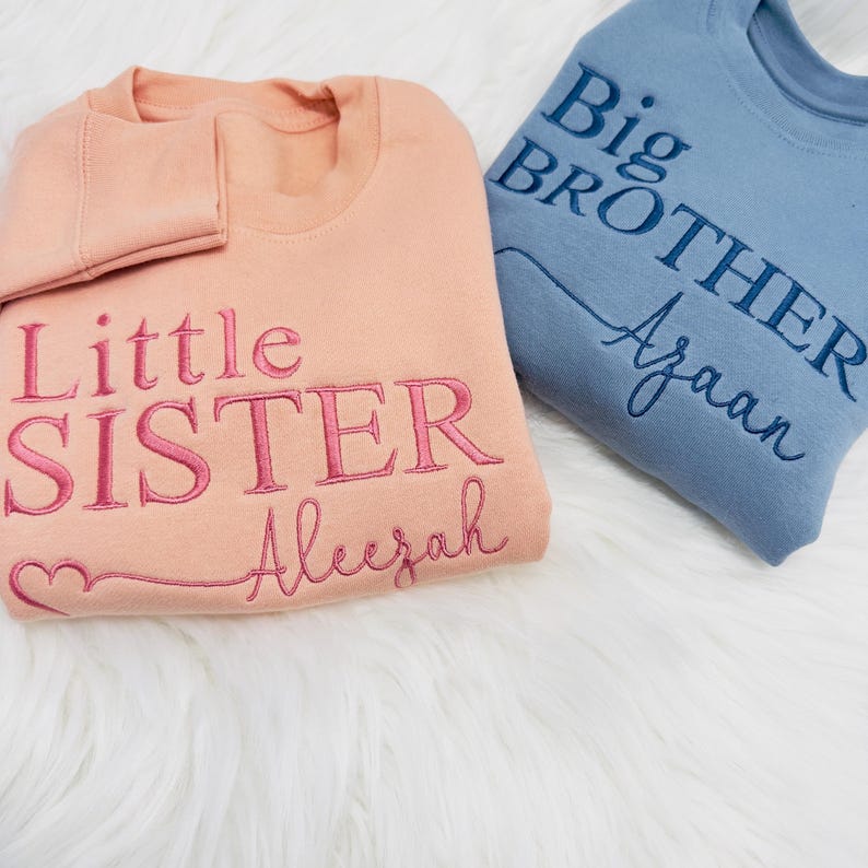 May include: Two sweatshirts, one peach and one blue, with embroidered text. The peach sweatshirt says "Little Sister Aleezah" with a pink heart. The blue sweatshirt says "Big Brother Ayaan".