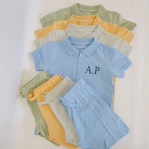 May include: A set of light blue, yellow, and green clothing for a child. The set includes a short-sleeved polo shirt with a zipper and the letters "A.P." embroidered on the chest, and a pair of matching shorts.