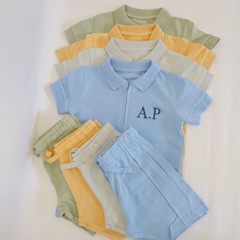 May include: A set of light blue, yellow, and green clothing for a child. The set includes a short-sleeved polo shirt with a zipper and the letters "A.P." embroidered on the chest, and a pair of matching shorts.