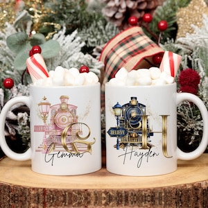 May include: Two white ceramic mugs with train designs, filled with marshmallows and candy canes. One mug features a pink train with the name "Gemma" and a gold "G". The other mug has a blue train with the name "Hayden" and a gold "H".