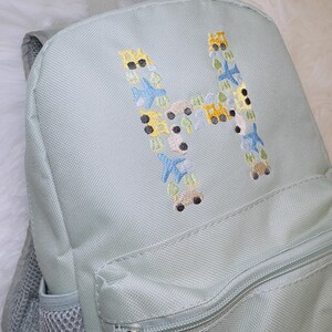May include: A light green backpack with a zippered pocket and a mesh pocket. The front of the backpack features an embroidered letter "H" made up of small, colorful vehicles and airplanes.