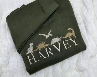 May include: A dark green sweatshirt with a white embroidered dinosaur design and the name "HARVEY" in white.