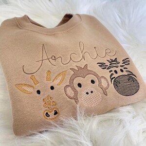 May include: A tan sweatshirt with embroidered animal faces, including a giraffe, monkey, and zebra. The name "Archie" is embroidered above the animals.