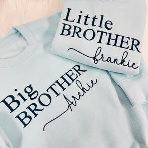 May include: Two light blue sweatshirts with black embroidered text. One sweatshirt says "Big Brother Archie" and the other says "Little Brother Frankie".