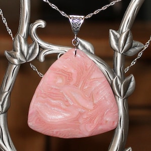 May include: A silver necklace with a triangular, pink pendant. The pendant has a swirling pattern of lighter pink and white. The necklace is displayed on a decorative silver stand with leaf accents.