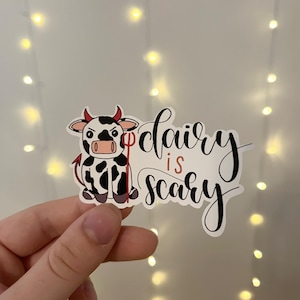 May include: A white and black cow with red horns and a red tail is holding a red pitchfork. The cow is looking at the camera with a menacing expression. The text "dairy is scary" is written in black cursive lettering.