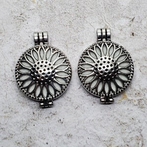 Puede incluir: Dos colgantes de locket de metal plateado con un diseño de girasol de esmalte blanco. Los lockets tienen una abertura con bisagra y un diseño de filigrana decorativo.