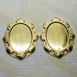 May include: Two gold-toned metal oval frames with ornate details. The frames are blank and ready for a photo or other embellishment.