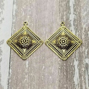 May include: Two gold-toned metal pendant charms with a diamond shape and intricate floral designs. The charms have a loop at the top for attaching to jewelry.