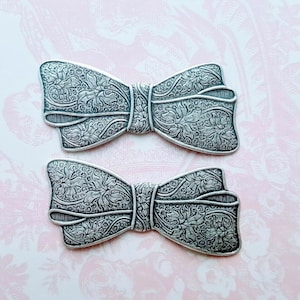 May include: Two silver-toned metal bow tie shapes with an intricate floral design. The bows are slightly different sizes and have a vintage look.