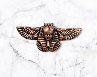 May include: A copper-colored metal pin with a detailed design of an Egyptian pharaoh's head with wings. The pharaoh's head is wearing a crown and has a beard. The wings are spread out on either side of the head.