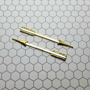 May include: Two gold-coloured arrow-shaped metal charms with a textured design. The charms are on a white background.