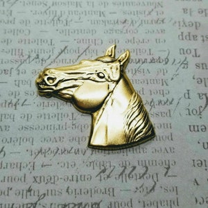 May include: A gold-colored metal horse head figurine. The horse's head is facing to the right. The figurine is detailed with a mane and a textured surface.