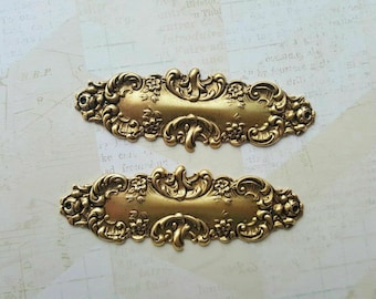 May include: Two ornate gold-toned metal embellishments with a decorative floral design. The embellishments are shaped like elongated ovals with a raised center section.
