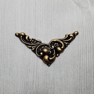 May include: Ornate, antique-style corner bracket in a bronze finish. The decorative metal piece features a symmetrical design with swirling patterns, floral accents, and a vintage aesthetic. Ideal for furniture restoration or craft projects.