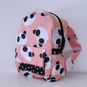 Puede incluir: Una mini mochila rosa con una cremallera negra y un bolsillo de lunares negros y blancos. La mochila está cubierta con un patrón de caras de panda negras y blancas.