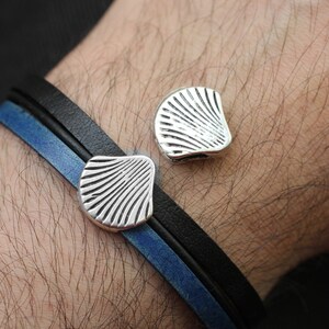 Op de afbeelding: Een leren armband met twee zilveren schelpvormige bedels. De armband is zwart en blauw.