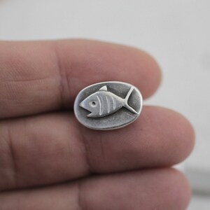 以下が含まれることがあります： 魚の形をした銀製のチャーム。チャームは楕円形で、ヒレとウロコのある魚の細かなデザインが施されています。