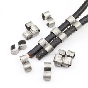 May include: A close-up of silver-toned metal cord ends for leather cord. The cord ends are designed to be crimped onto the ends of leather cords to create a finished look. The cord ends are scattered on a white surface.