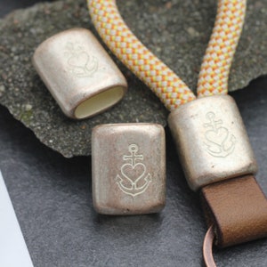 Puede incluir: Tres llaveros de metal plateados con un diseño de ancla y corazón grabado. Los llaveros están unidos a una cuerda amarilla y blanca y una correa de cuero marrón.
