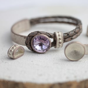 Könnte beinhalten: Ein geflochtenes Lederarmband in hellbraun und weiß. Das Armband ist mit einem violetten Kristall in einer silbernen Fassung und dekorativen silbernen Perlen verziert.