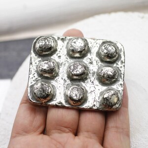 May include: A silver-toned metal rectangular object with nine raised, rounded bumps. The metal has a textured, aged appearance, with dark areas contrasting against the lighter silver. The object is held in a hand.