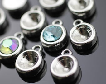 May include: A collection of silver-toned metal settings for jewelry making. The settings are round and have a raised lip around the edge. Some of the settings have clear glass stones in them, while others are empty. The stones are clear, blue, and iridescent green.