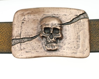 May include: A copper-colored belt buckle with a skull design. The buckle has a cracked surface and a detailed skull with empty eye sockets.