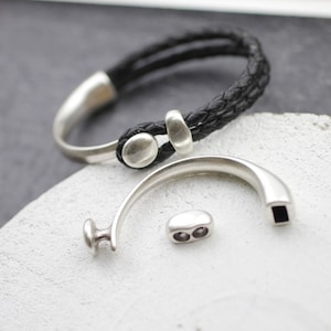 Puede incluir: Un brazalete de cuero negro con cierres de metal plateado y un anillo de metal plateado con una abertura cuadrada.