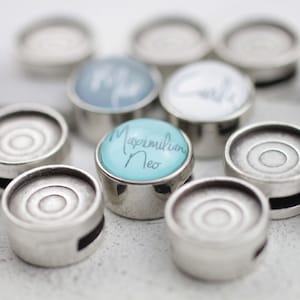 May include: A close-up of a set of silver charm beads with a blue and white charm bead that says "Maximilian Neo".