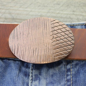 Könnte beinhalten: Eine kupferfarbene ovale Gürtelschnalle mit strukturierter Oberfläche und einem braunen Ledergürtel. Die Schnalle hat ein einzigartiges Design mit vertikalen Linien auf der einen Seite und einem Rautenmuster auf der anderen. Die Schnalle ist an einer blauen Jeans befestigt.