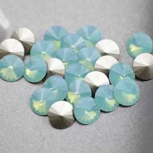 May include: A group of light blue iridescent glass rhinestones with silver backs. The rhinestones are scattered on a gray surface.